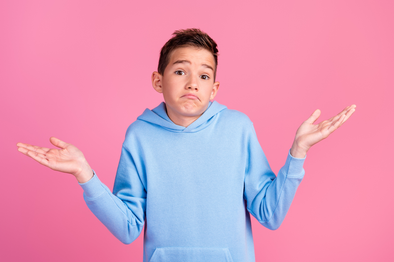 Vaaleansinipaitainen teinipoika kohauttaa hämmentyneenä olkiaan ja levittelee käsiään pinkkiä taustaa vasten.