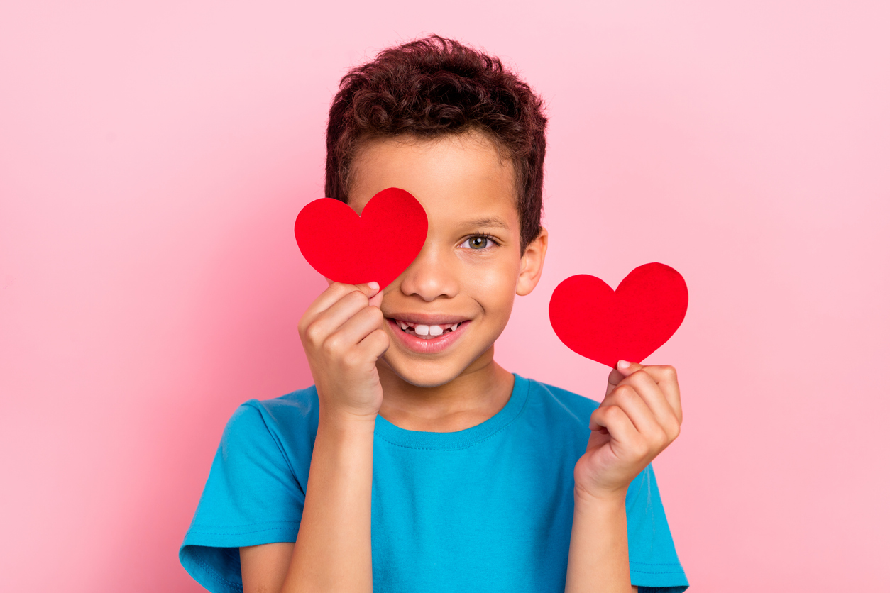 Tumma poika pitää kahta paperista sydäntä käsissään vaaleanpunaista taustaa vasten.