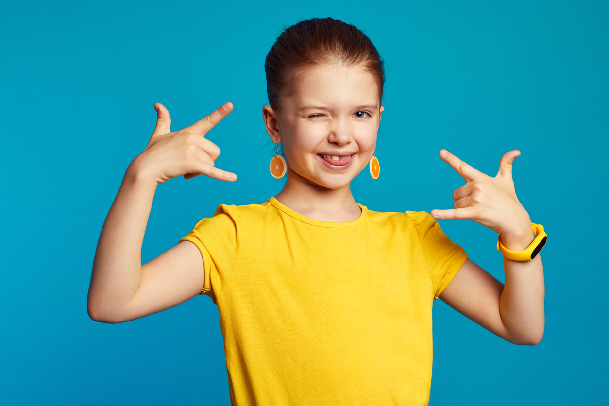 Pieni tyttö näyttää molemmilla käsillään käsimerkkejä ja iskee silmää turkoosia taustaa vasten.