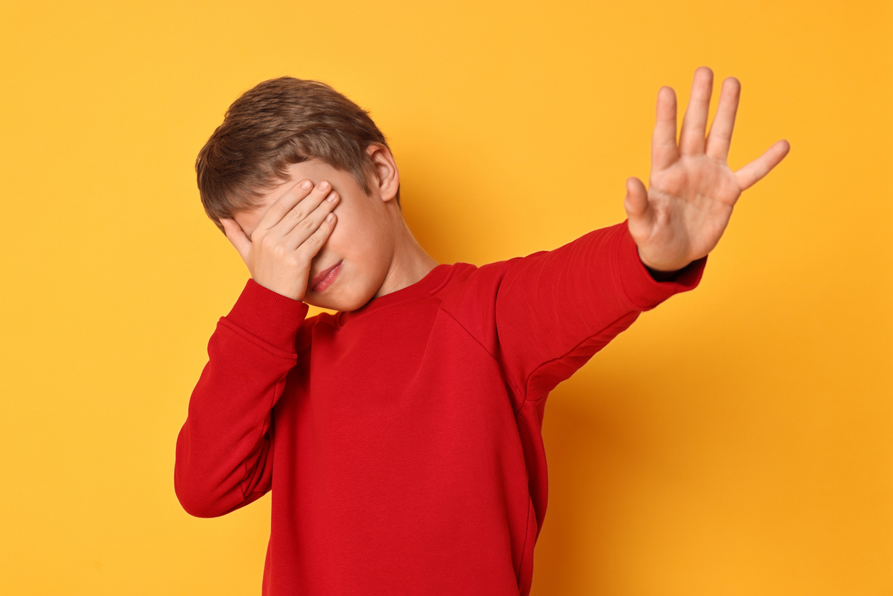 Poika punaisessa paidassa peittää silmänsä ja pitää torjuvaa kättä edessään keltaista taustaa vasten.