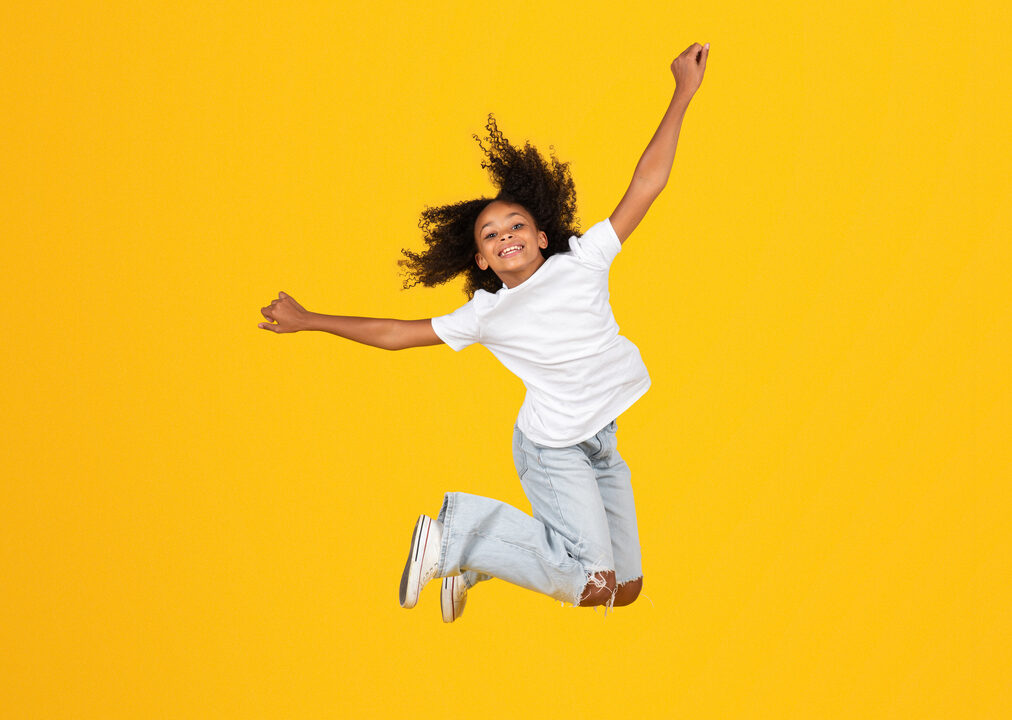 Tumma lapsi hyppää korkealle ilmaan keltaista taustaa vasten.