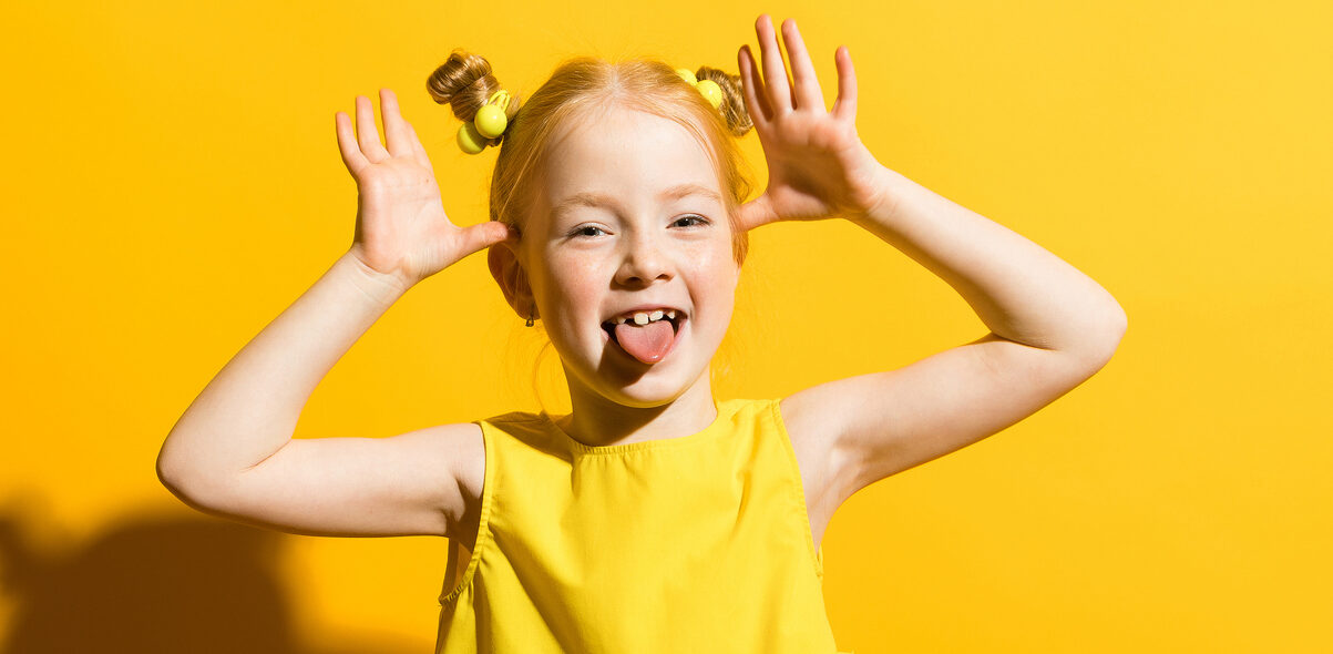 Saparopäinen tyttö ilmeilee ja näyttää kieltä kameralle keltaista taustaa vasten.